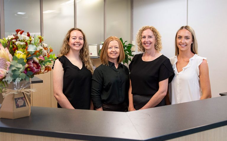 Four female Sackett and Weir staff members smiling standing behind front desk